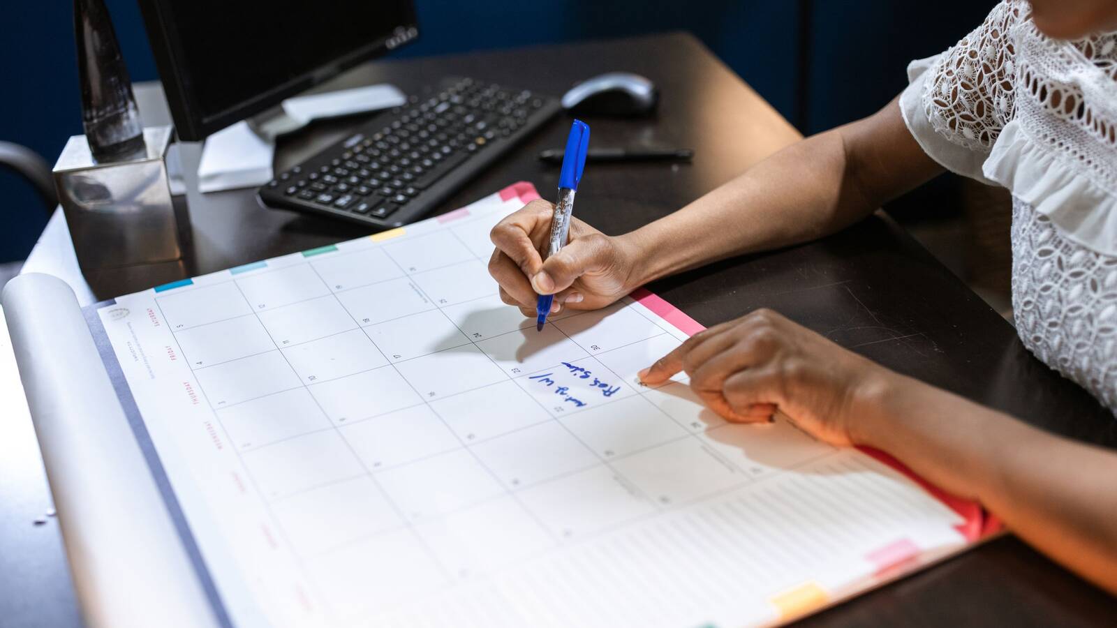 A person writing a note with a blue pen on a large monthly desk calendar