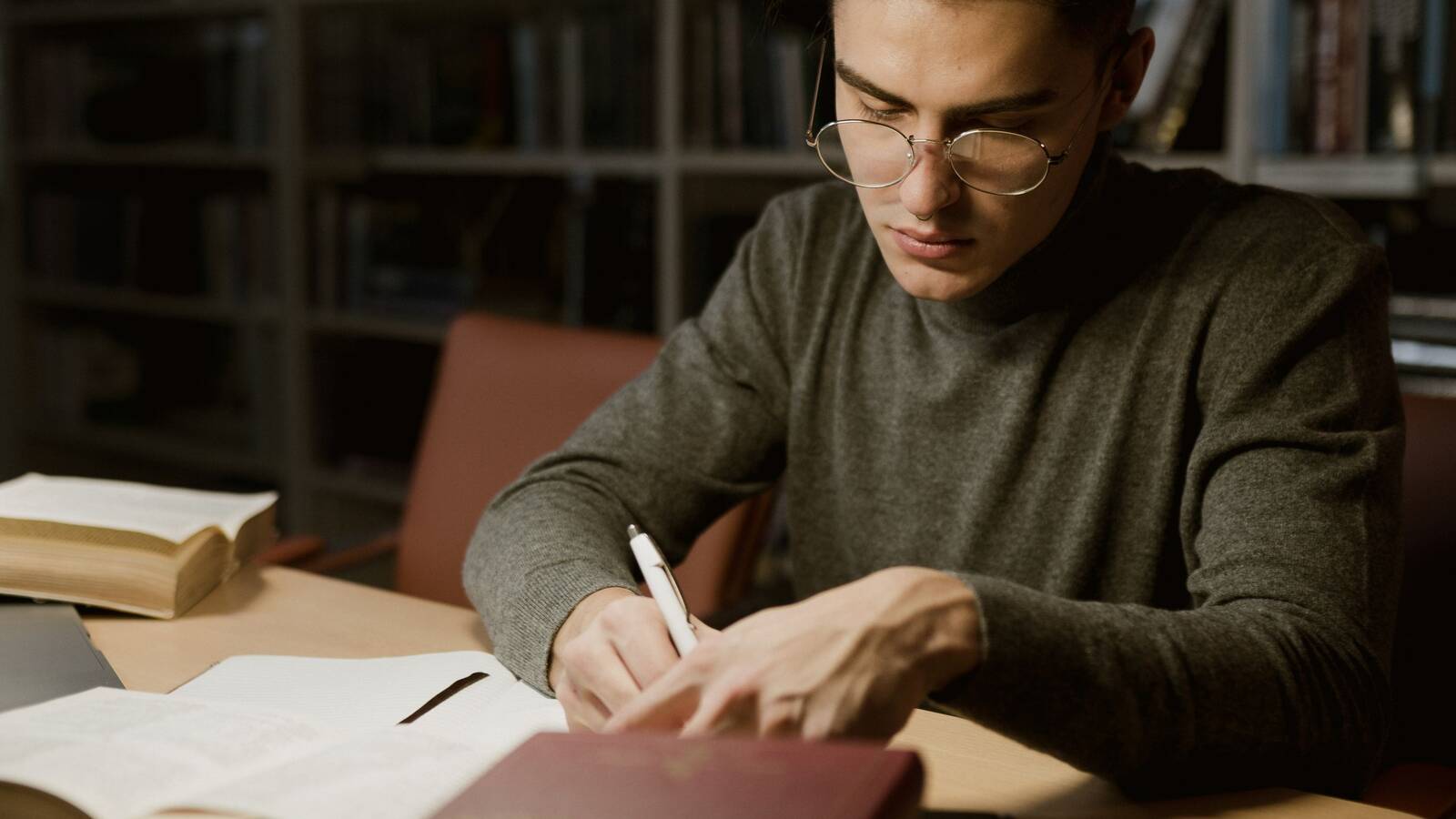 A man in glasses writing notes at a library desk, with open books in front of him and shelves behind