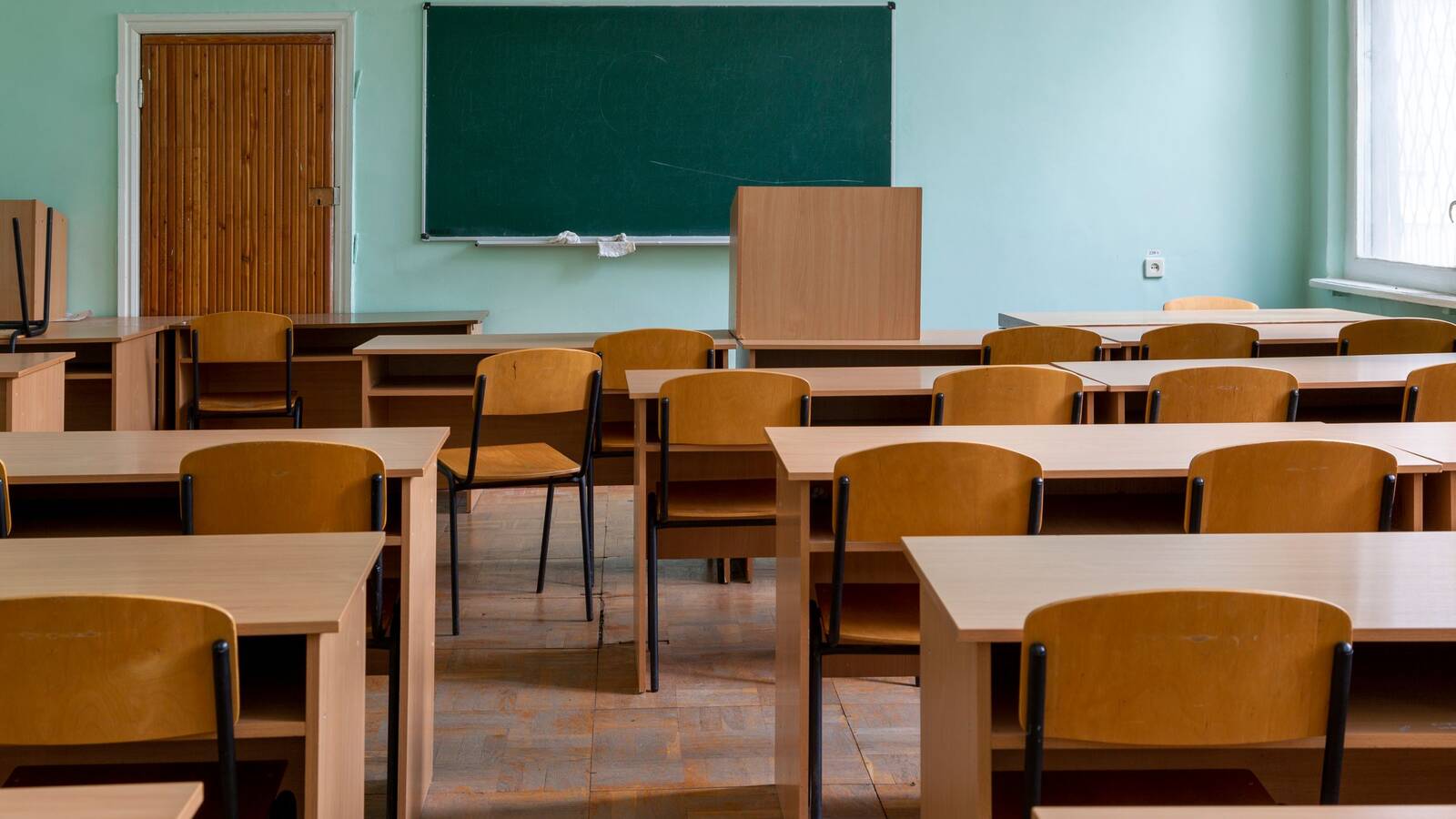 An empty classroom with rows of wooden desks and chairs in front of a green chalkboard