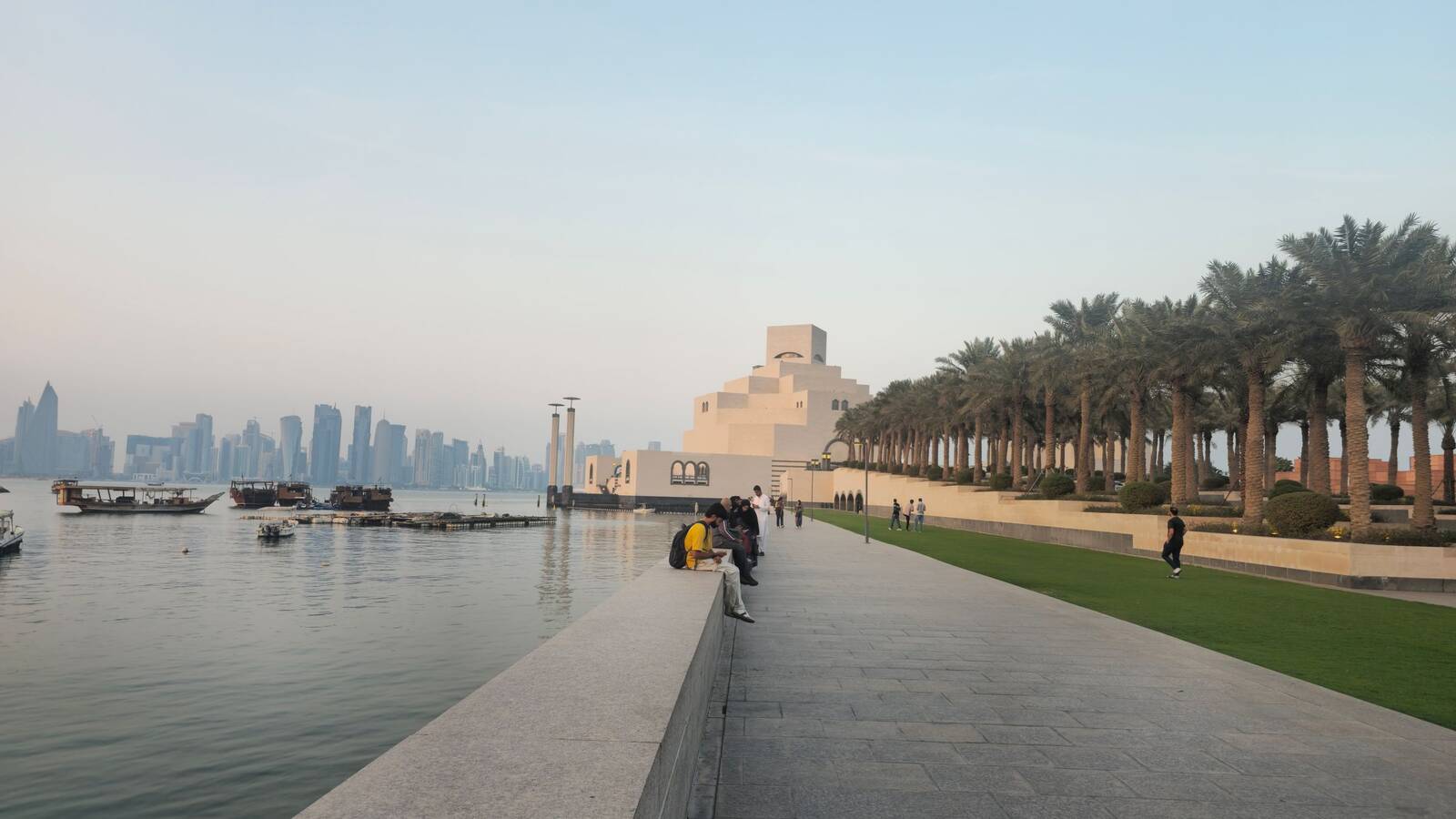 A palm-lined waterfront path in Doha, with the Museum of Islamic Art and the city towers across the bay