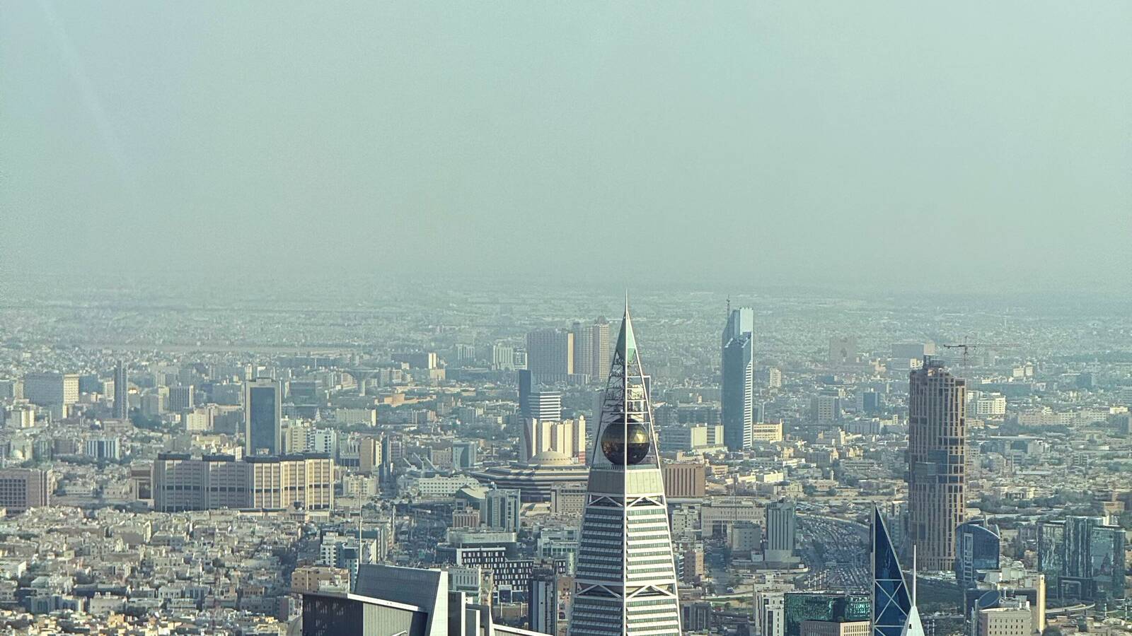 Aerial view of Riyadh on a hazy day, with the pointed Al Faisaliah Tower in front of the city