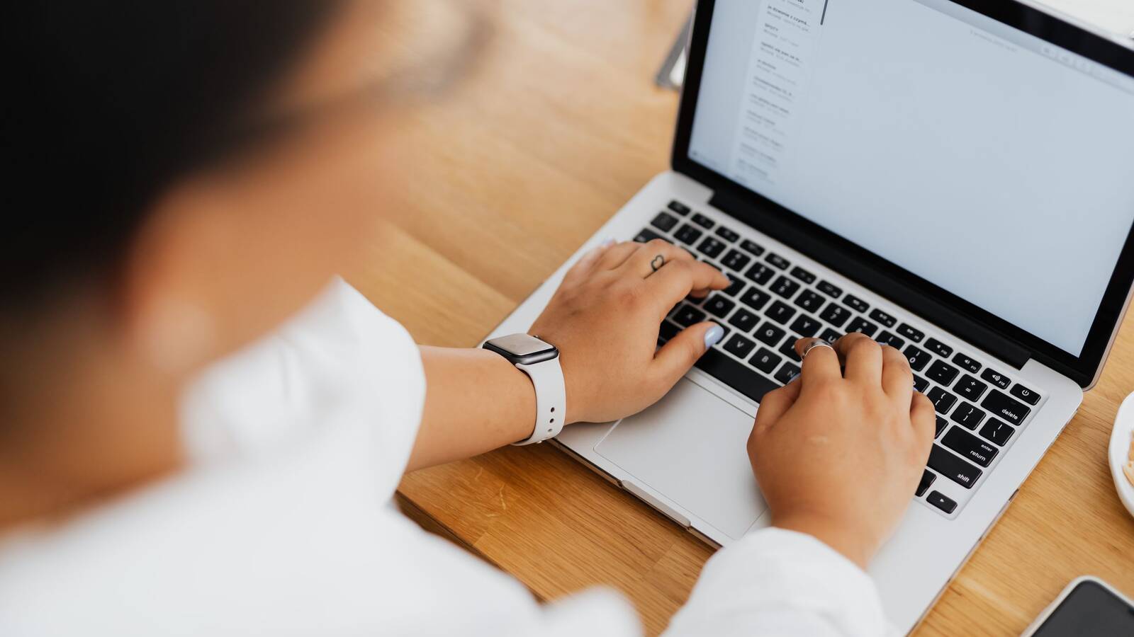A person typing on a laptop at a wooden desk, seen from over the shoulder