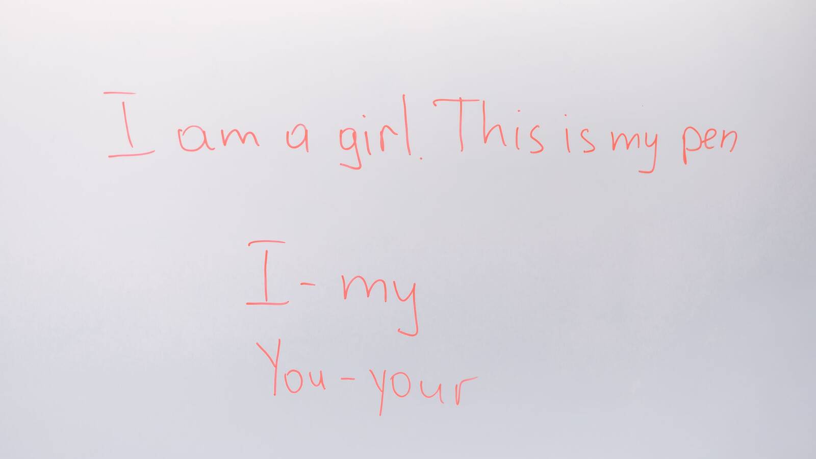 An English lesson handwritten in red on a whiteboard: 'I am a girl. This is my pen', 'I - my' and 'You - your'